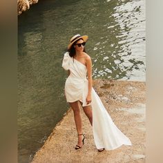 a woman in a white dress and hat standing next to the water with her hand on her hip