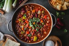 a large pot filled with chili and beans next to bread, vegetables and crackers