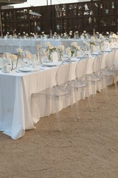 a long table is set up with clear chairs and white linens for an outdoor wedding reception