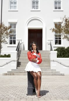 a woman in a red jacket and white skirt posing for the camera with her hand on her chin