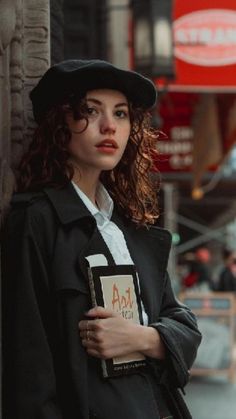 a woman in a black coat and hat leaning against a wall with her hand on her hip