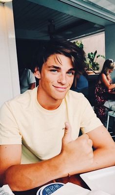 a young man sitting at a table giving the thumbs up sign