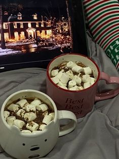 two mugs of hot chocolate and marshmallows on a bed next to a christmas pillow