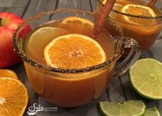 two mugs filled with orange juice and cinnamon on a table next to sliced apples