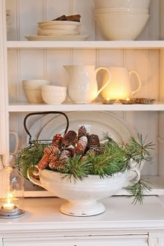 a white bowl filled with pine cones on top of a counter next to candles and dishes