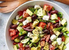 a white bowl filled with cucumber, tomato and olive salad next to wooden spoons