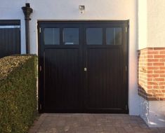 a black garage door on the side of a white house with brick walkway and potted plants