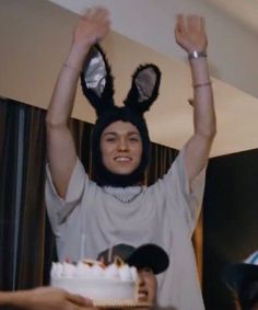 a woman in bunny ears holds her hands up above her head while holding a cake with candles on it