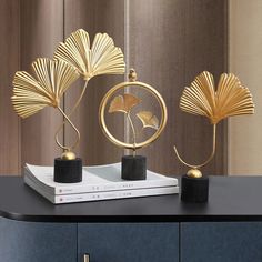 three golden sculptures on top of a black table next to a white book and brown wall