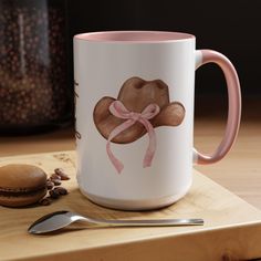 a coffee mug with a pink ribbon on it next to a spoon and cupcake