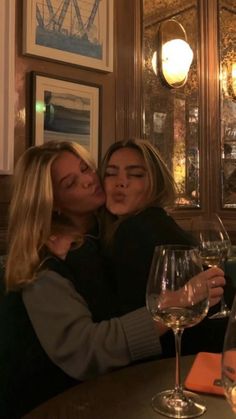 two women sitting at a table with wine glasses in front of them and one is kissing the other