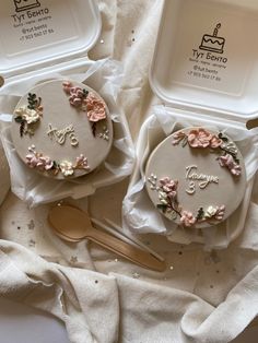 two cakes decorated with flowers are sitting in plastic containers on a white cloth covered table