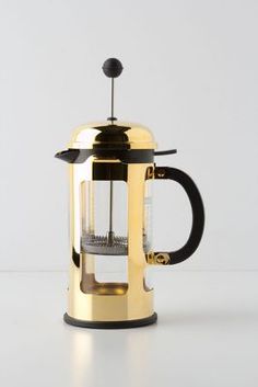 a gold colored coffee pot with a glass in the top and handle on it, sitting next to a white wall