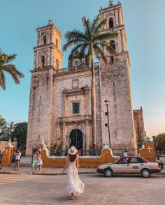 a woman in a white dress and hat walking towards an old church with palm trees