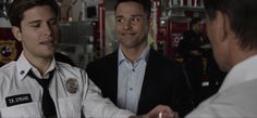 two men shaking hands while standing in front of a firetruck with another man looking on