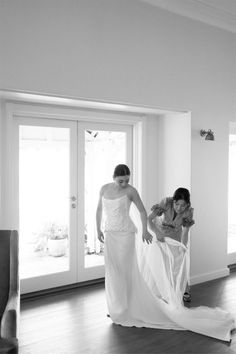 two women in dresses are walking through a room with open doors and one woman is holding the other's dress