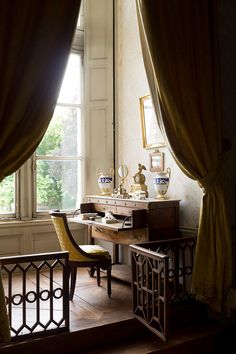 a desk and chair in front of a window with drapes on the windowsill