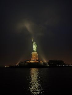 the statue of liberty is lit up at night with its lights on and reflecting in the water