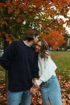 a man and woman standing next to each other in front of trees with leaves on the ground