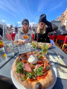 two women sitting at an outdoor table with pizza and drinks in front of their faces