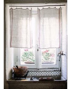 a kitchen window with two white curtains and a bowl on the counter in front of it