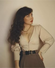 a woman with long hair wearing a white shirt and brown skirt, standing against a wall