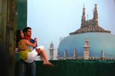 a man holding a woman in his arms on top of a roof with buildings behind him