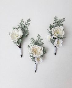 three boutonnieres with white flowers and greenery on the ends are shown