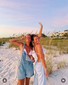 two women standing on the beach with their arms in the air and one woman wearing overalls