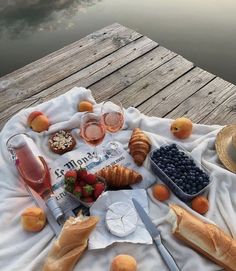 an assortment of food is laid out on a picnic blanket near the water, along with fruit and bread