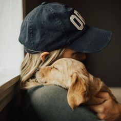 a woman holding a dog wearing a baseball cap on top of her head while sitting next to a window