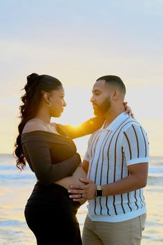 a man and woman standing next to each other in front of the ocean at sunset