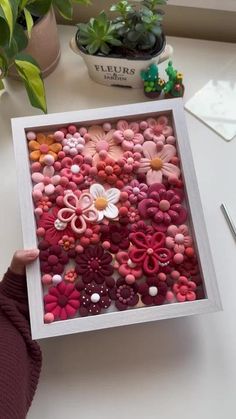 a person holding up a box filled with buttons and flower shapes in front of a potted plant