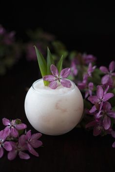 a white vase filled with purple flowers on top of a wooden table next to greenery