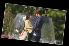 two people standing in front of a water fountain with trees and bushes behind them, one holding the other's back