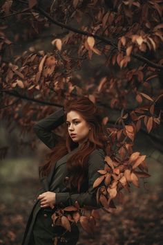 a woman with long red hair standing in front of a leafy tree and looking at the camera