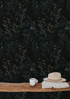 a hat, cup and saucer sit on a table in front of a floral wallpaper