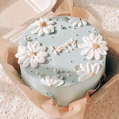 there is a blue cake with white flowers on it in a cardboard box next to lace doily