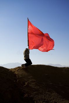 a person sitting on top of a hill holding a red flag in the air royalty photography