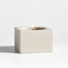 a small white square candle holder with a lit candle in it's center on a plain surface