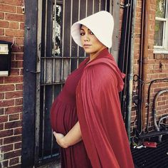 a pregnant woman in a red dress and white hat is standing outside an iron gate