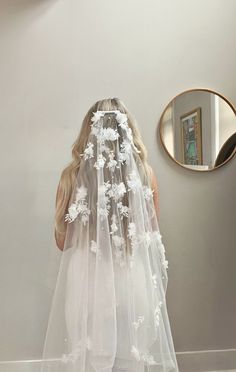 the back of a bride's veil with white flowers on it, standing in front of a mirror