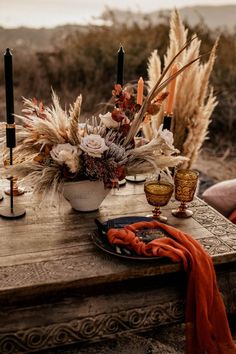 a wooden table topped with a vase filled with flowers and candles next to an orange scarf