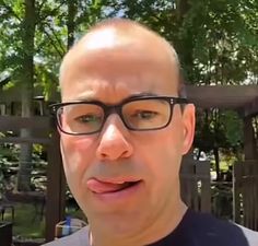 a bald man wearing glasses is making a funny face at the camera with trees in the background