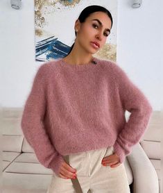 a woman standing in front of a white couch wearing a pink sweater and beige pants