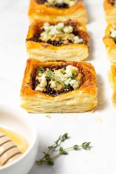 several small pastry items on a white surface next to a bowl of honey and herbs