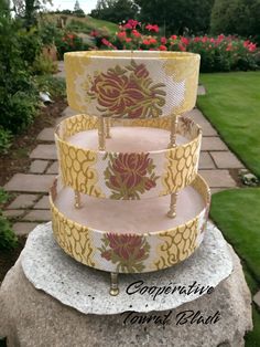 three tiered cake stand sitting on top of a rock in front of some flowers