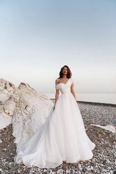 a woman in a white wedding dress standing on rocks near the ocean with her arms behind her back