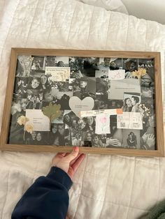 a person holding up a framed photo with pictures and papers attached to it on top of a bed