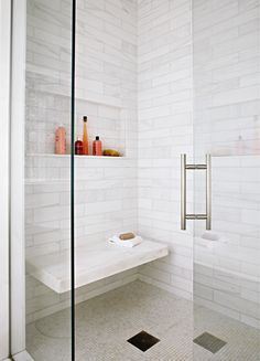 a white tiled bathroom with glass shower doors and shelves on the wall, along with toiletries